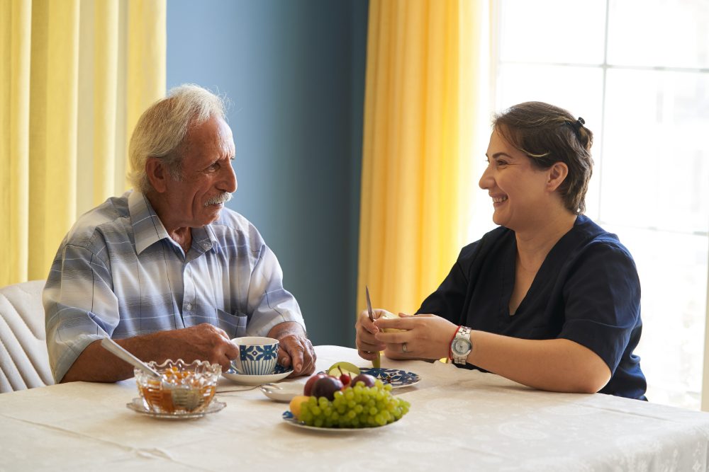 An older man talking with a visitor at his kitchen table